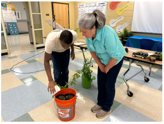 Key Center Goes Green for Earth Week - student and teacher looking at the bucket of soil, student watering the plant.png