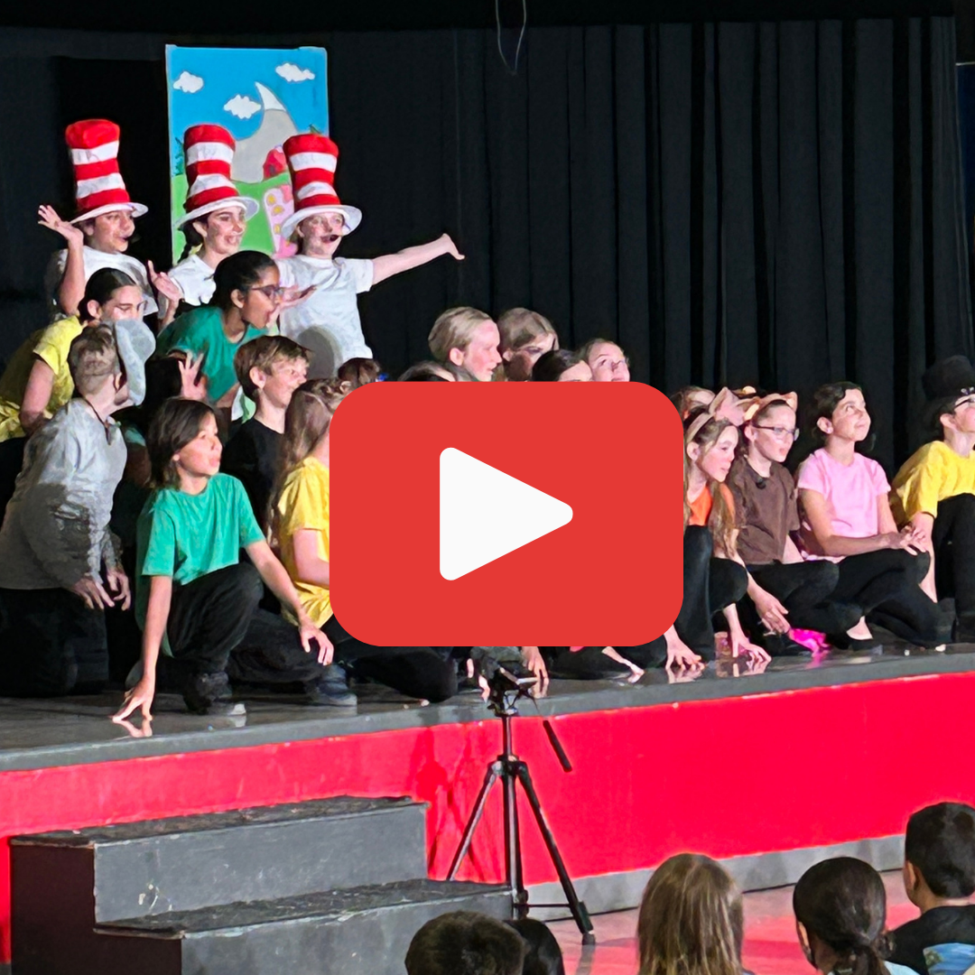 A group of students performs on stage in colorful costumes, with three wearing tall red and white striped hats. A red play button overlayed