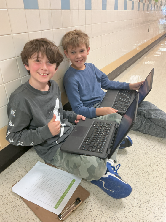 Two smiling young students sit on a school hallway floor working together on laptops.