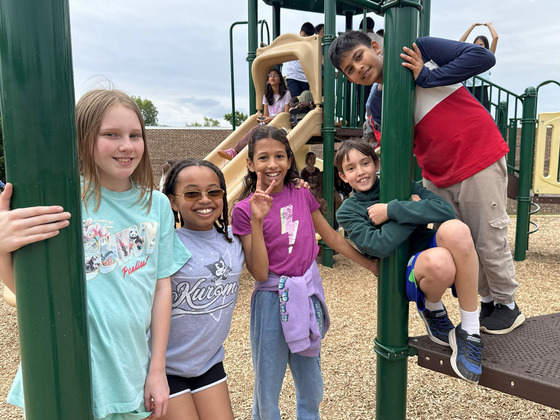 Five smiling, elementary-aged students pose together on a green and tan playground structure during recess.