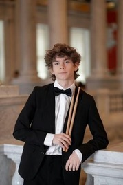 A male student wearing a dress suit with a bowtie poses in front of a marble railing while holding drumsticks