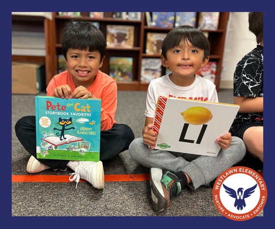 Two students sit on the carpet and hold a library book while smiling at the camera.