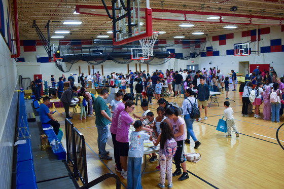 Wide shot of gymnasium showing several booths with over 100 young kids running in between them.