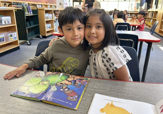 2 preK students sitting at a library table with a book in front of them.