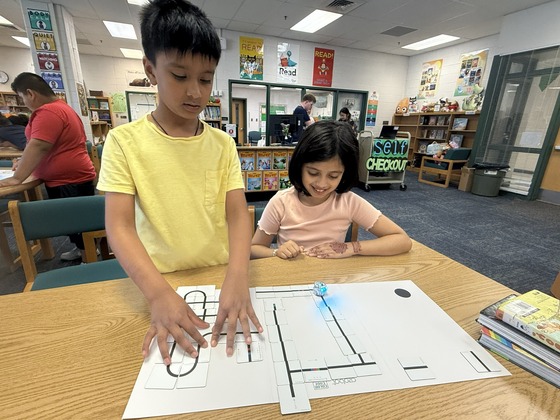 Two young students work together at a library table, interacting with an Ozobot robot on a printed coding track map.