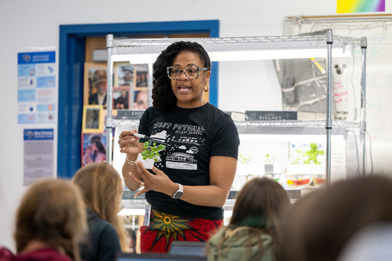 A teacher holds a plant while talking to a classroom of students