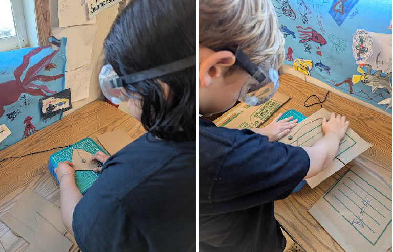 Two students wearing goggles using cardboard cutters.  Read text below image for a deeper description.