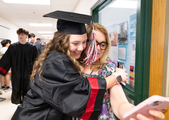 A student in graduation gown and hat hugs a teacher who is taking a selfie. 