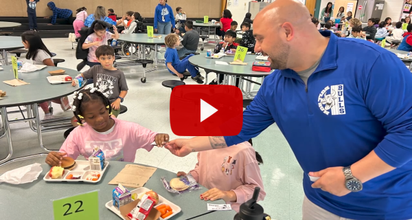 A screenshot of a video: An adult hands a sticker to a student in a cafeteria during lunch. 