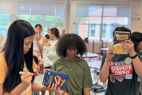 Students examine and label pasta boxes during a hands-on classroom activity, with several students collaborating around cafeteria tables.