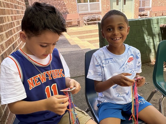 Two elementary school students sit outside weaving colorful yarn crafts on small pieces of cardboard.