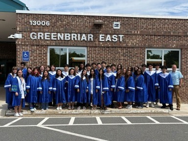 A large group of high school graduates in blue caps and gowns poses outside the brick entrance of Greenbriar East school.