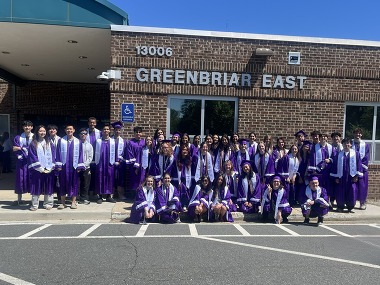 A large group of high school graduates in purple caps and gowns poses outside the brick entrance of Greenbriar East school.
