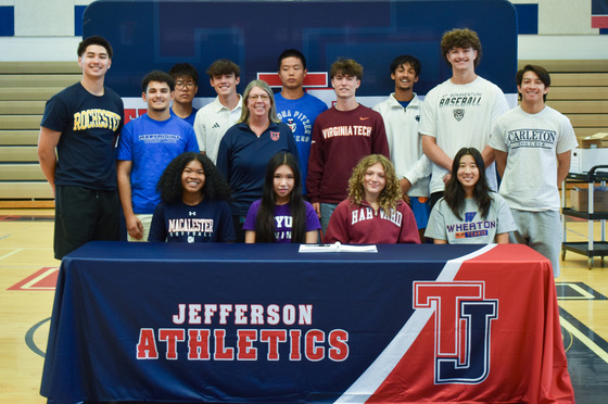 13 of the 14 Jefferson Next Level Athletes pose behind a table in two rows in front of a TJ Athletics backdrop