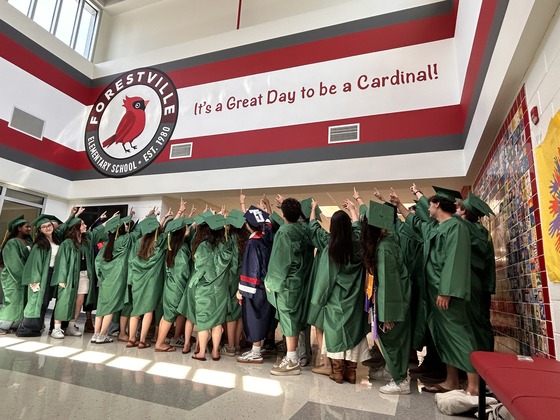 Graduating seniors in caps and gowns return to Forestville Elementary posing beneath the school’s cardinal logo and motto.