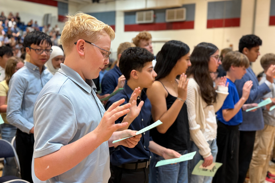 Seventh grade students stand in a gymnasium with hands raised as they recite the National Junior Honor Society pledge during an NJHS pinning ceremony.