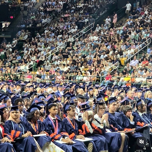 Students in cap and gowns as family looks on from stands