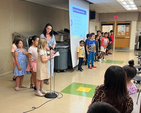 Students stand with their teacher and take turns using the microphone to address the audience.