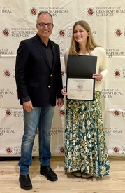 A female student poses with a certificate in front of a flat Univerity of Maryland college style backdrop
