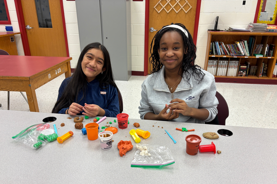 Two students smile while shaping colorful modeling clay at a classroom table covered with clay tools and small creations.
