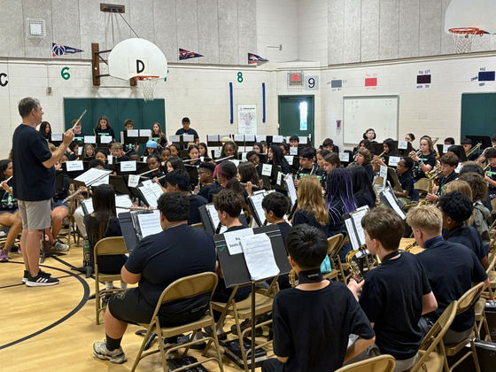 Student band members perform in a school gym as their conductor leads the ensemble during a daytime concert. (99 characters)