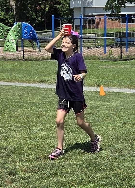 A soggy student smiling and carrying a cup of water on her head for the water bucket relay during field day.