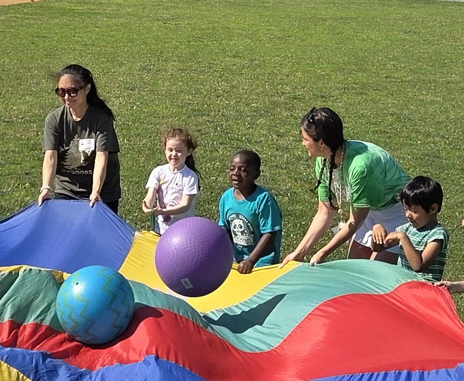 Students and staff bouncing playground balls on top of a rainbow parachute while enjoying field day.