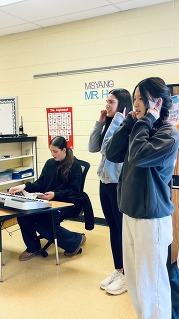 Two women stand holding their ears while another sits playing an electronic keyboard in a classroom