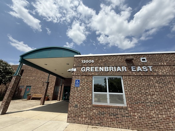 The front entrance of Greenbriar East Elementary School with the school name displayed on a brick building beneath a blue sky with clouds.
