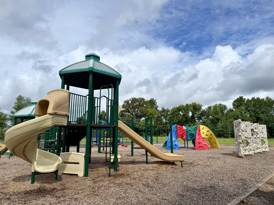 Green and tan playground equipment, colorful climbing structures, and a rock wall beneath a cloudy sky.