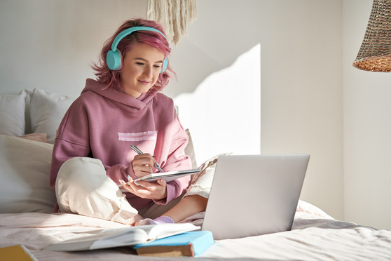 Girl sitting on her bed with headphones on while writing on a notebook and looking at her laptop