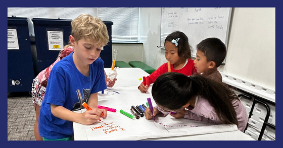 Multiple students use markers to write about their favorite part of the summer reading book on a shared sheet of chart paper.