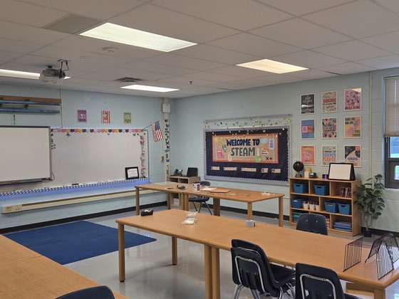 Brightly lit STEAM classroom set up with wooden worktables, whiteboards, and a 'Welcome to STEAM' bulletin board.
