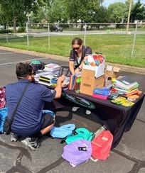 A family selects notebooks, folders, and other school supplies from a table while backpacks sit nearby.