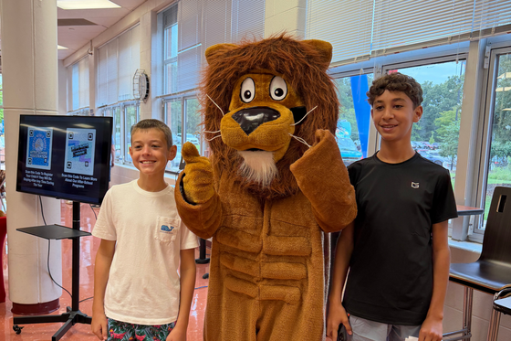 Two young students posing with the school lion mascot near a digital information display in a school hallway.