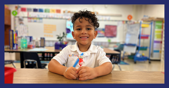 A student sits at a table and smiles at the camera. He holds a blue highlighter and a red lollipop.
