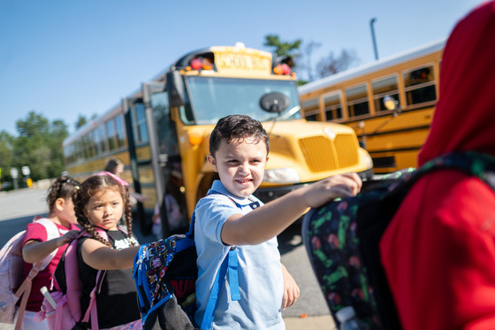Students walk from a bus to school holding each other's backpacks to stay in line