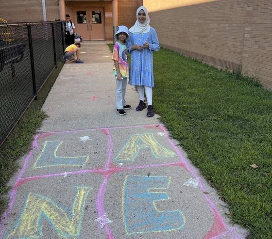Students standing in front of a chalk drawing of LANE on the sidewalk of Kiss and Ride loop