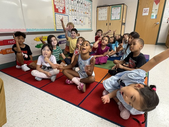 Kindergarten students sit together on a colorful classroom rug and eagerly raise their hands during a lesson.