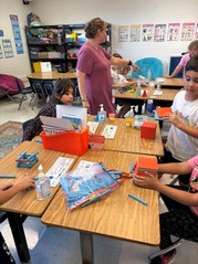 teacher walking past students sitting at desks, playing with math manipulatives like chains and hundreds blocks