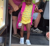 a girl in pink shirt with backpack and a badge hanging off. she is walking down the stairs of bus with an adult helping her down