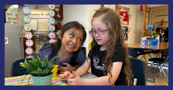 Two students sit at the table in the “house” section of the classroom.