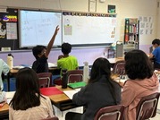 students raising hands sitting at desks. math problems on smart board in the background