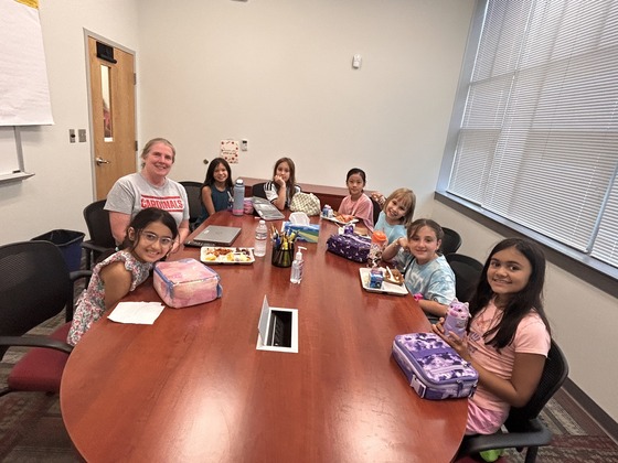 A group of students and the school principal sit around a conference table with their lunches, smiling at a camera