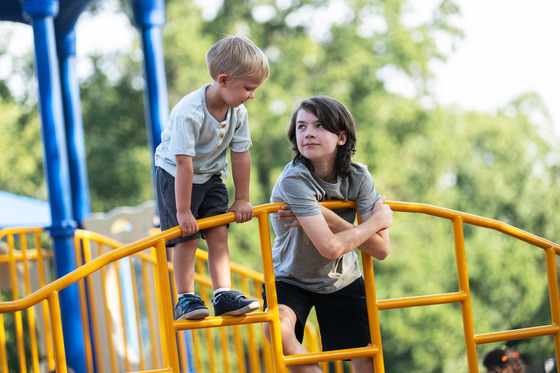 A 6th grader looks up at a kindergartener standing above him on a climbing structure on a playground.