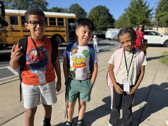 Three students wearing backpacks smile as they arrive at school, with a school bus behind them.
