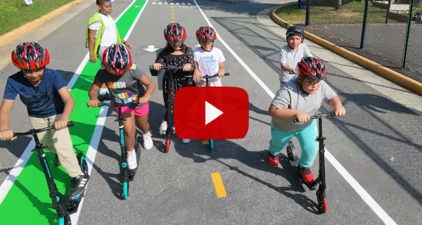 Video play button over a screenshot of students on push scooters.