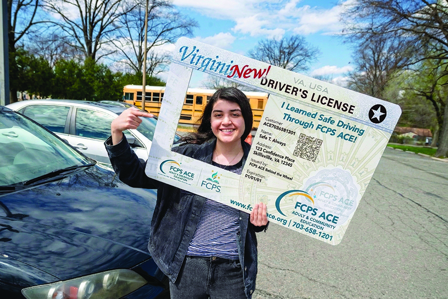 A smiling student in a parking lot looks through a hole cut in a large license where her photo would be.