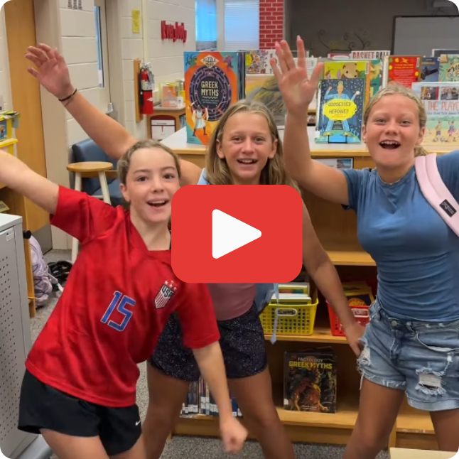 three smiling girls holding hands out wide standing in a library with a red and white play graphic overlay