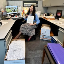 Ashlee Young sitting at her desk surrounded by boxes of paper files that are being sorted and archived.